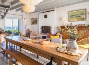 Een ruime woonkamer met een houten tafel, zitplaatsen, een open haard en uitzicht op de omgeving.