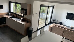 A modern kitchen with a large window, a dining area with a sofa, and a sliding glass door leading to an outdoor patio. - Vacation home in Pordic - close to the sea (Pordic)