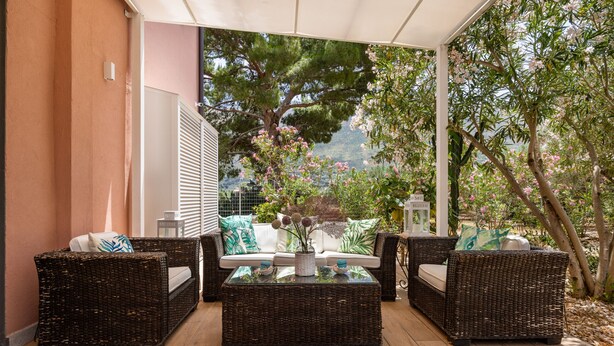A covered outdoor seating area with wicker furniture and a glass-top coffee table, overlooking a garden with trees and a view of the landscape. - Villa Giovanna con Piscina by Wonderful Italy (Castellammare del Golfo)