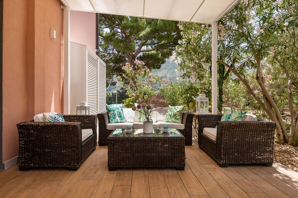 Un espace extérieur aménagé, abrité et doté de meubles en osier et d’une table basse en verre, offrant une vue sur un jardin arboré et sur le paysage environnant.