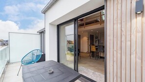 Ein Balkon mit Tisch und blauem Stuhl, eine Glasschiebetür, die zu einem Essbereich mit Stühlen und Tisch im Inneren führt.