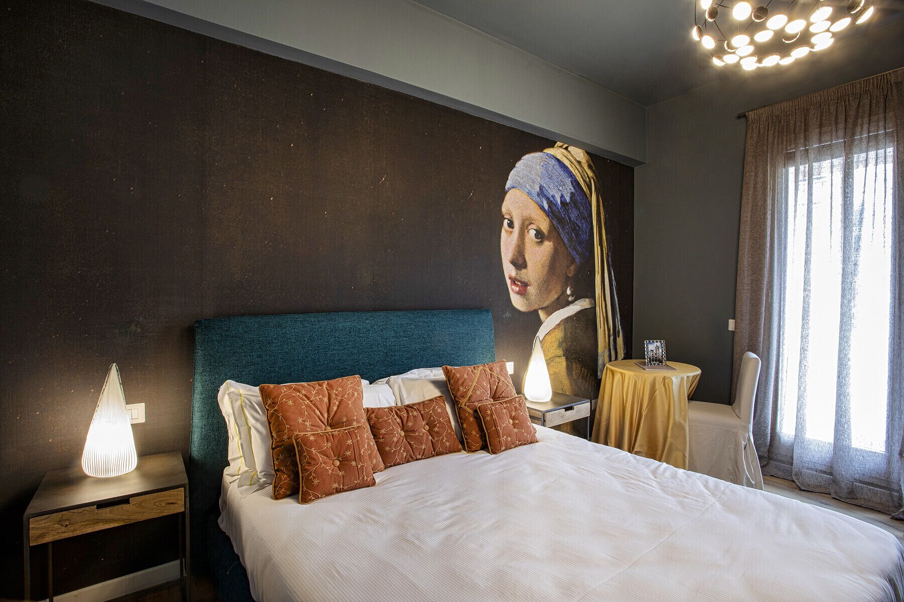 A bedroom with a large bed, a nightstand with a lamp, a table with a picture frame, and a wall with a large painting.