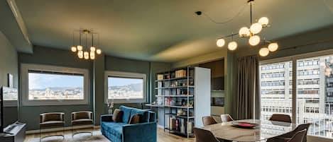 Un espacio habitable moderno con comedor, sofá azul, estantería y grandes ventanas con vistas a la ciudad.