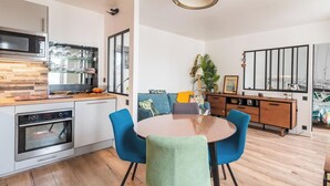 A modern kitchen with a built-in oven, a dining area with a round table and blue chairs, and a living space with a sofa and a large window. - L'Escale Paimpolaise 2 Stars - historic center (Paimpol)