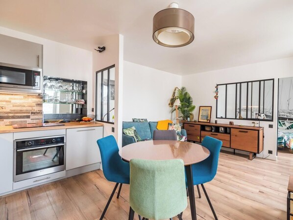 Une cuisine moderne dotée d’un four encastré, un coin repas avec une table ronde et des chaises bleues, et un espace de vie comprenant un canapé et une grande fenêtre.