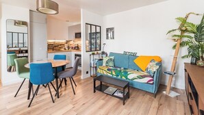 A modern living room with a sofa, coffee table, and chairs. There is a dining table with chairs, a kitchen area, and a large plant. - L'Escale Paimpolaise 2 Stars - historic center (Paimpol)