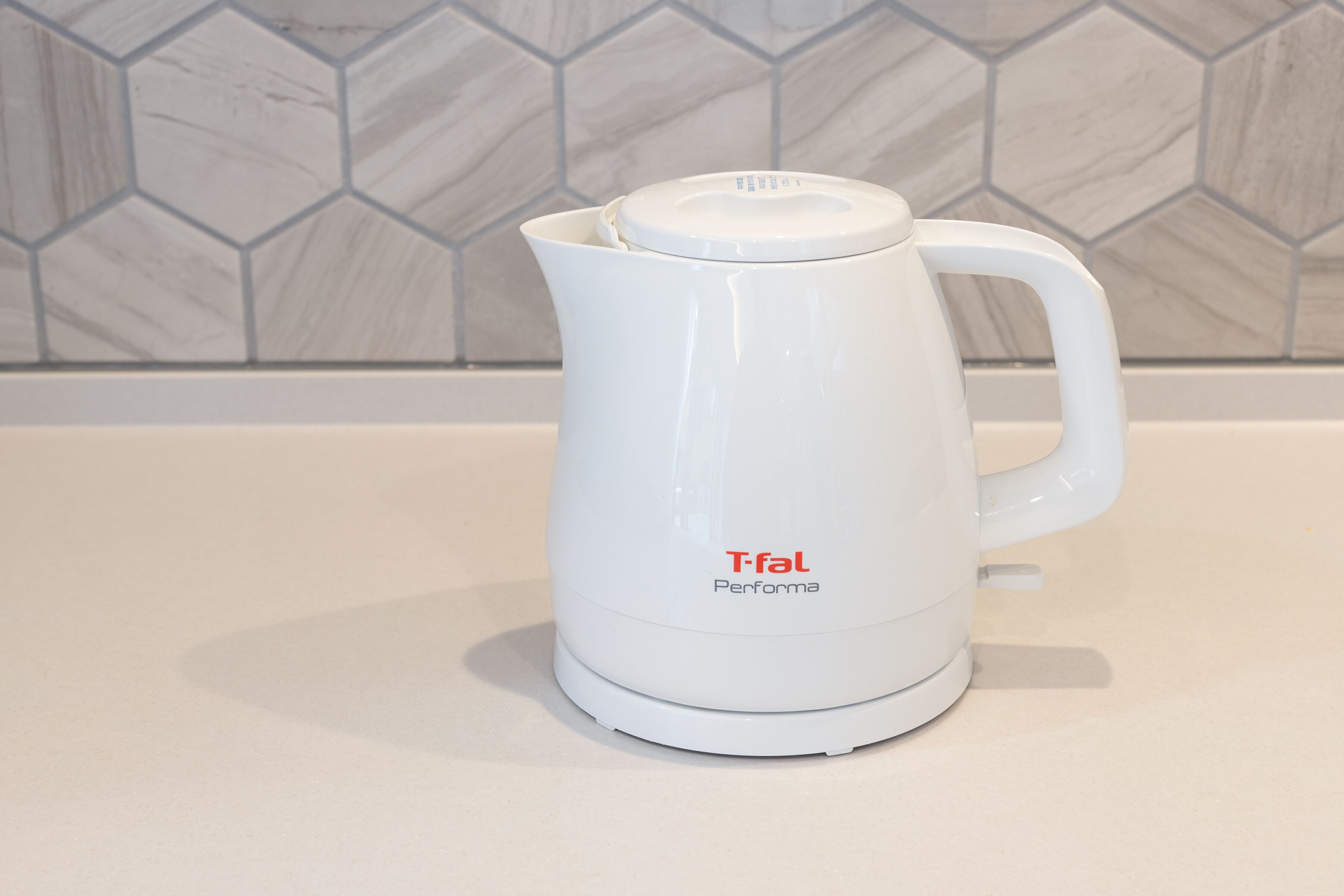 Ένα λευκό ηλεκτρικό βραστήρι Tefal Performa πάνω σε έναν λευκό πάγκο, με ένα εξαγωνικό πλακάκι ως διακοσμητικό στοιχείο στον τοίχο.