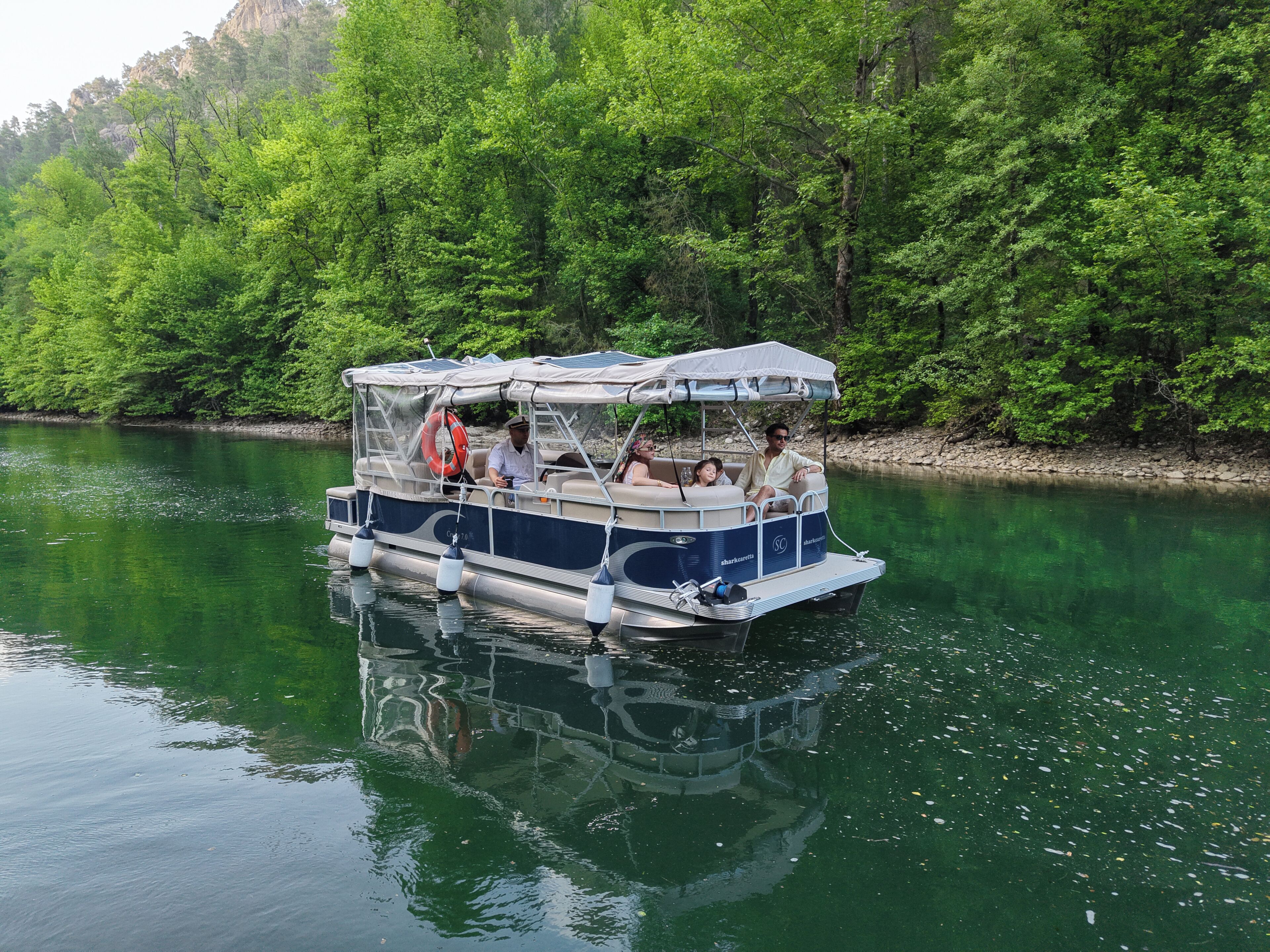 A boat with a covered seating area, passengers, and a river surrounded by greenery.