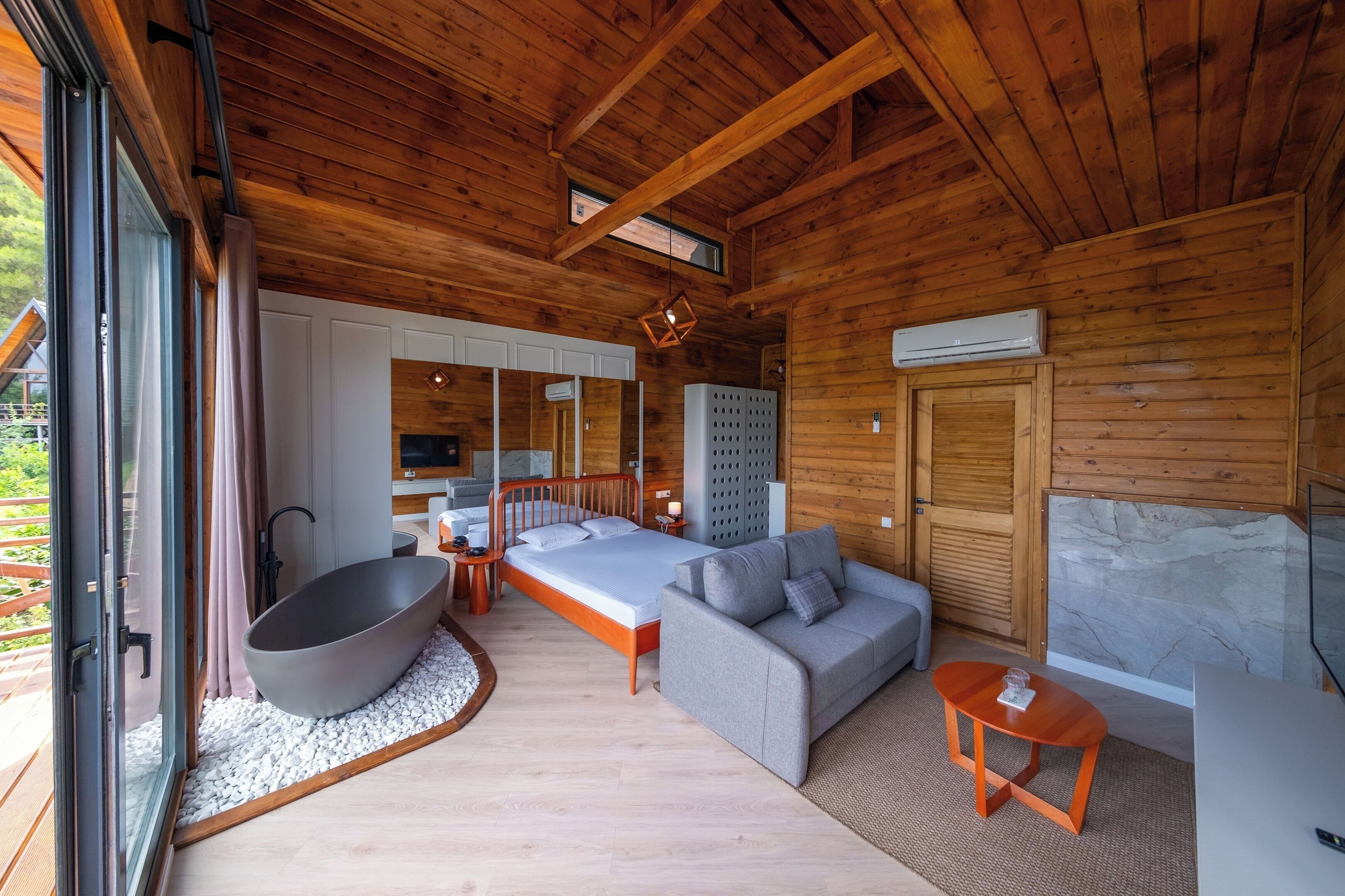 Un dormitorio de cabaña de troncos moderno con una cama grande, un sofá gris, una mesa de madera y una bañera espaciosa.