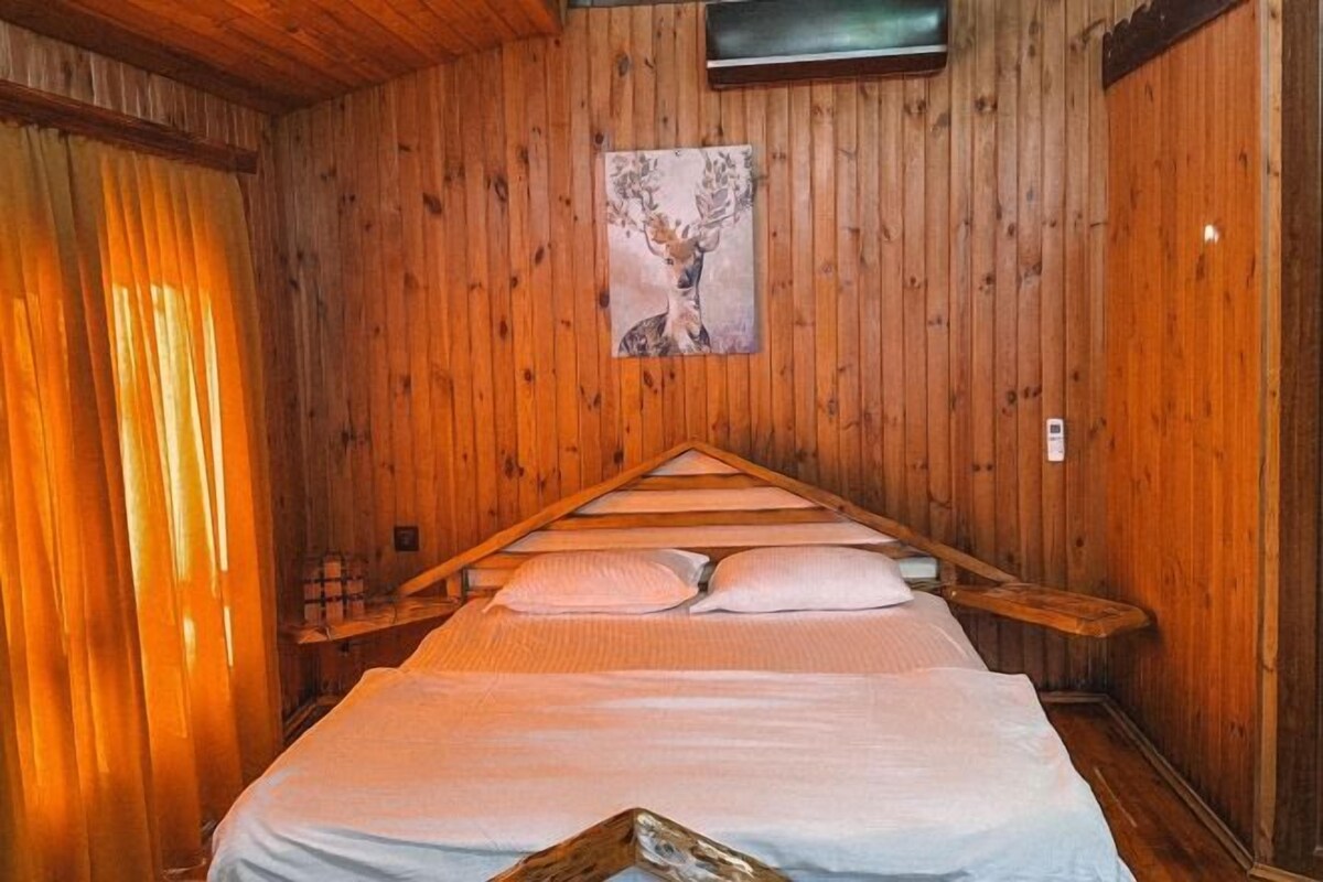A wooden cabin room with a bed, a painting of a deer, and a window with curtains.