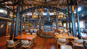 A rustic wooden interior with high ceilings, exposed beams, and a central wood-burning stove. Tables and chairs are arranged around the stove, and hanging light fixtures are visible. - Botanik Gol Evleri (Bucak)