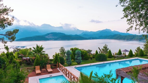 A swimming pool with a wooden deck and lounge chairs, surrounded by lush greenery and a view of a lake and mountains. - Botanik Gol Evleri (Bucak)
