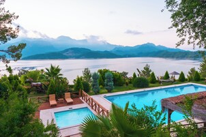 Una piscina con muelle de madera y sillas de descanso, rodeada de abundante vegetación y vistas a un lago y montañas.