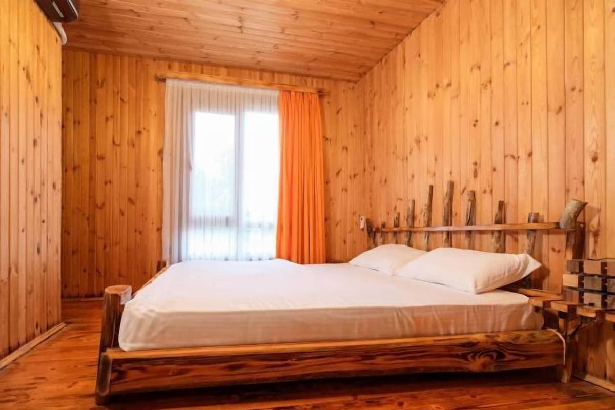 Quarto com painéis de madeira, uma cama, uma janela com cortinas e cabeceira de madeira.