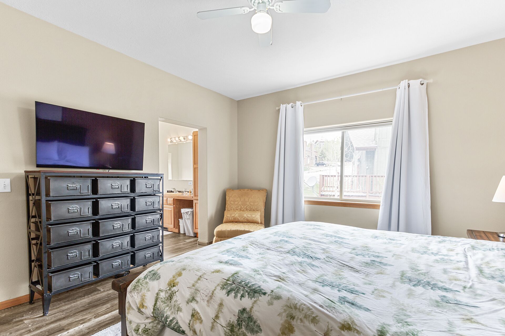 Una habitación de hotel con cama, cómoda con cajones, televisor, silla, ventana con cortinas y ventilador de techo.