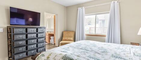 Una habitación de hotel con cama, cómoda con cajones, televisor, silla, ventana con cortinas y ventilador de techo.