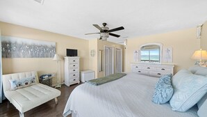 A bedroom with a bed, a dresser, a vanity, a television, a ceiling fan, and a large window with a view.