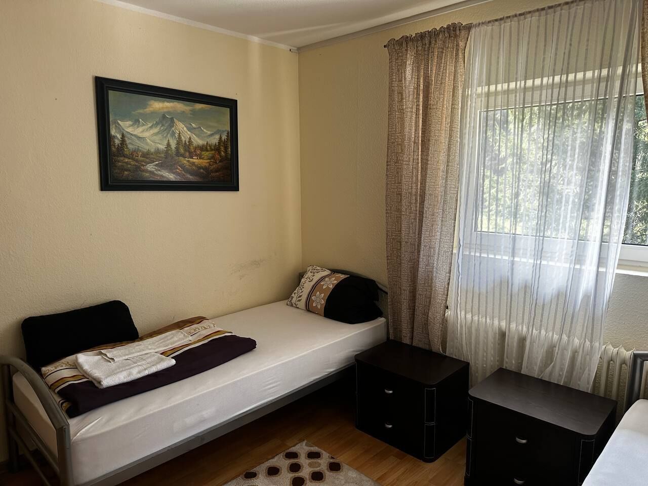 A single bed with a blanket, a bedside table, a small cabinet, a painting on the wall, and a window with curtains.