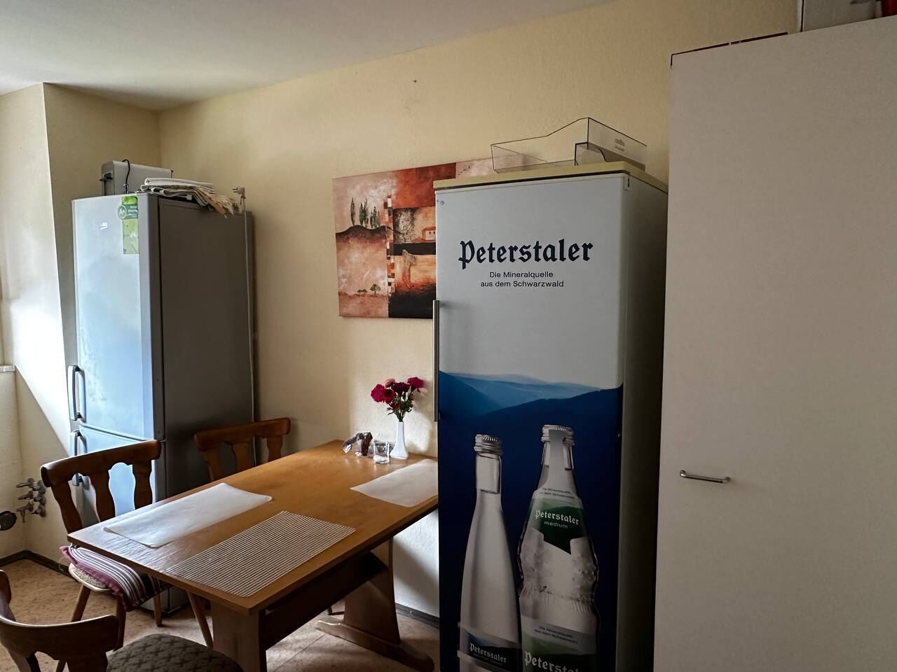 A dining area with a wooden table, chairs, a refrigerator, and a poster of Peterstaler mineral water.
