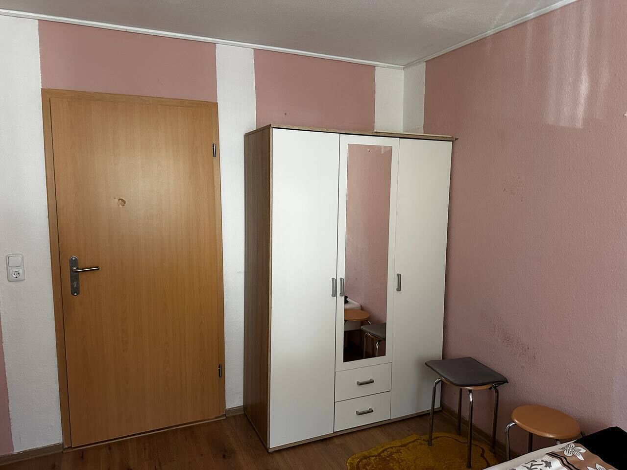 Ein Zimmer mit Holztür, einem weißen Kleiderschrank mit Spiegel, einem kleinen Hocker und einem Tisch mit gemustertem Tischlauf.