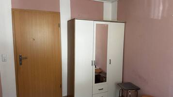 Ein Zimmer mit Holztür, einem weißen Kleiderschrank mit Spiegel, einem kleinen Hocker und einem Tisch mit gemustertem Tischlauf.
