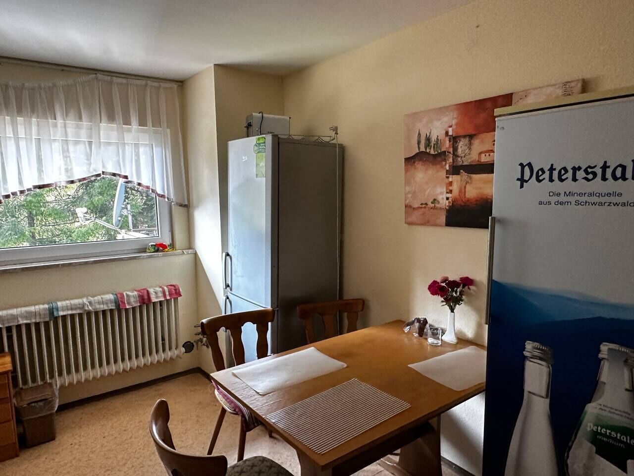 Ein kleiner Raum mit einem Holztisch, Stühlen, einer Heizung, einem Fenster mit Vorhängen, einem Kühlschrank und einem Poster an der Wand.
