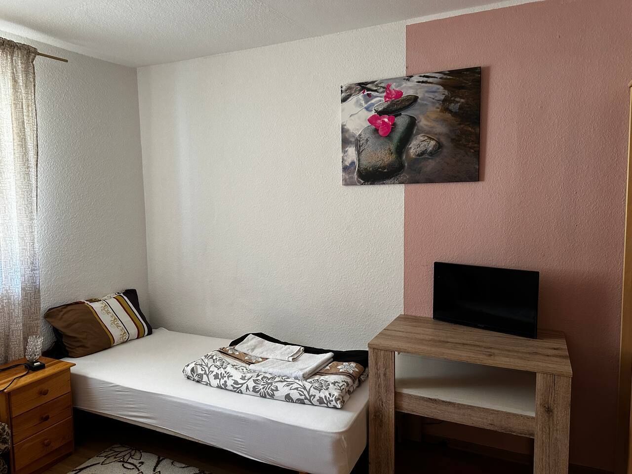 A bedroom with a bed, a wooden nightstand, a bedside table, a TV on a wooden stand, and a painting on the wall.