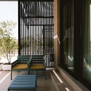 A balcony with two green chairs and a bench, a brick wall, and a view of the outside.