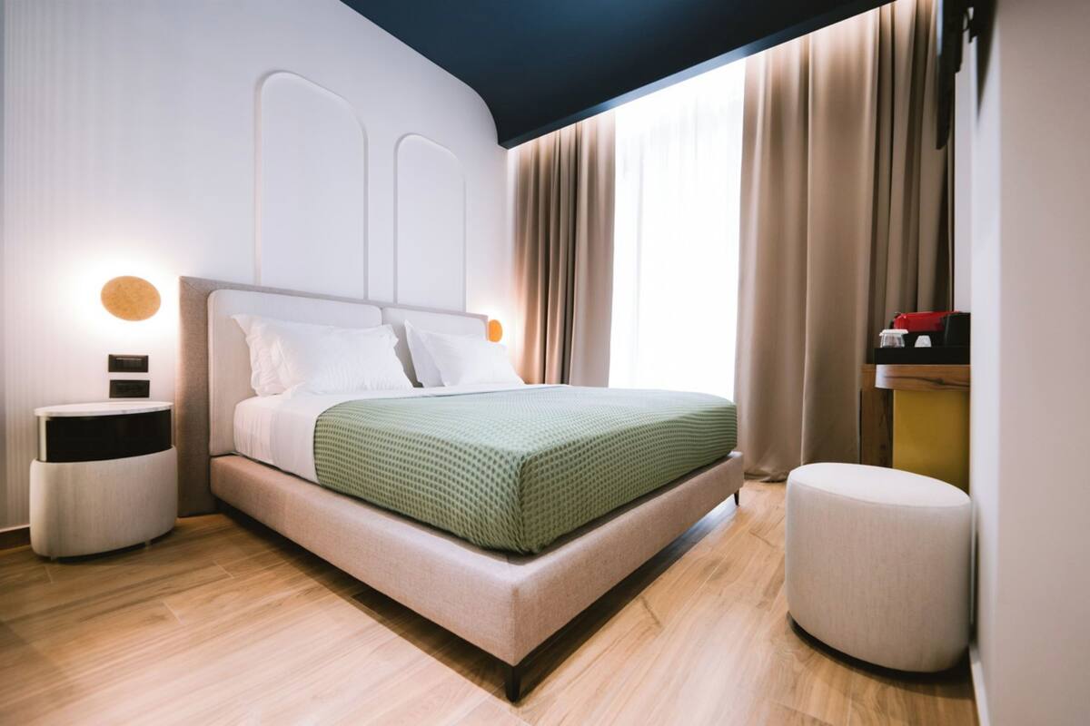A modern bedroom with a bed, bedside tables, a round ottoman, and a view of the outdoors.