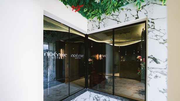 A hotel entrance with glass doors, a marble floor, and a wall with a green plant and flowers. - Hotel Noemie (Himarë)