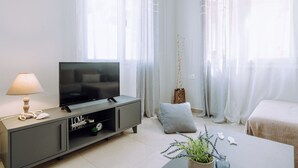Ett modernt vardagsrum med en platt-TV på en grå TV-bänk, ett bord med en växt och en säng med ett vitt överkast.