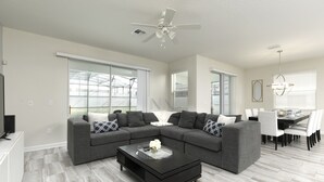 A modern living room with a sectional sofa, a coffee table, and a dining area with a table and chairs. - Luxury 6BR Townhome with Private Pool, Game Room & Resort Access Near Disney (Kissimmee)