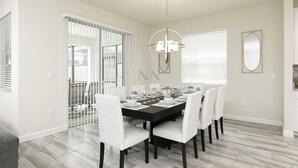 A dining area with a black table set for six, white chairs, a chandelier, and a large window with blinds. - Luxury 6BR Townhome with Private Pool, Game Room & Resort Access Near Disney (Kissimmee)