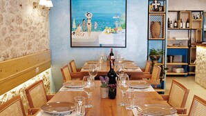 A dining area with a long wooden table set for a meal, surrounded by wicker chairs. A large painting of a beach scene is on the wall behind the table. - Himara 28 (Himarë)
