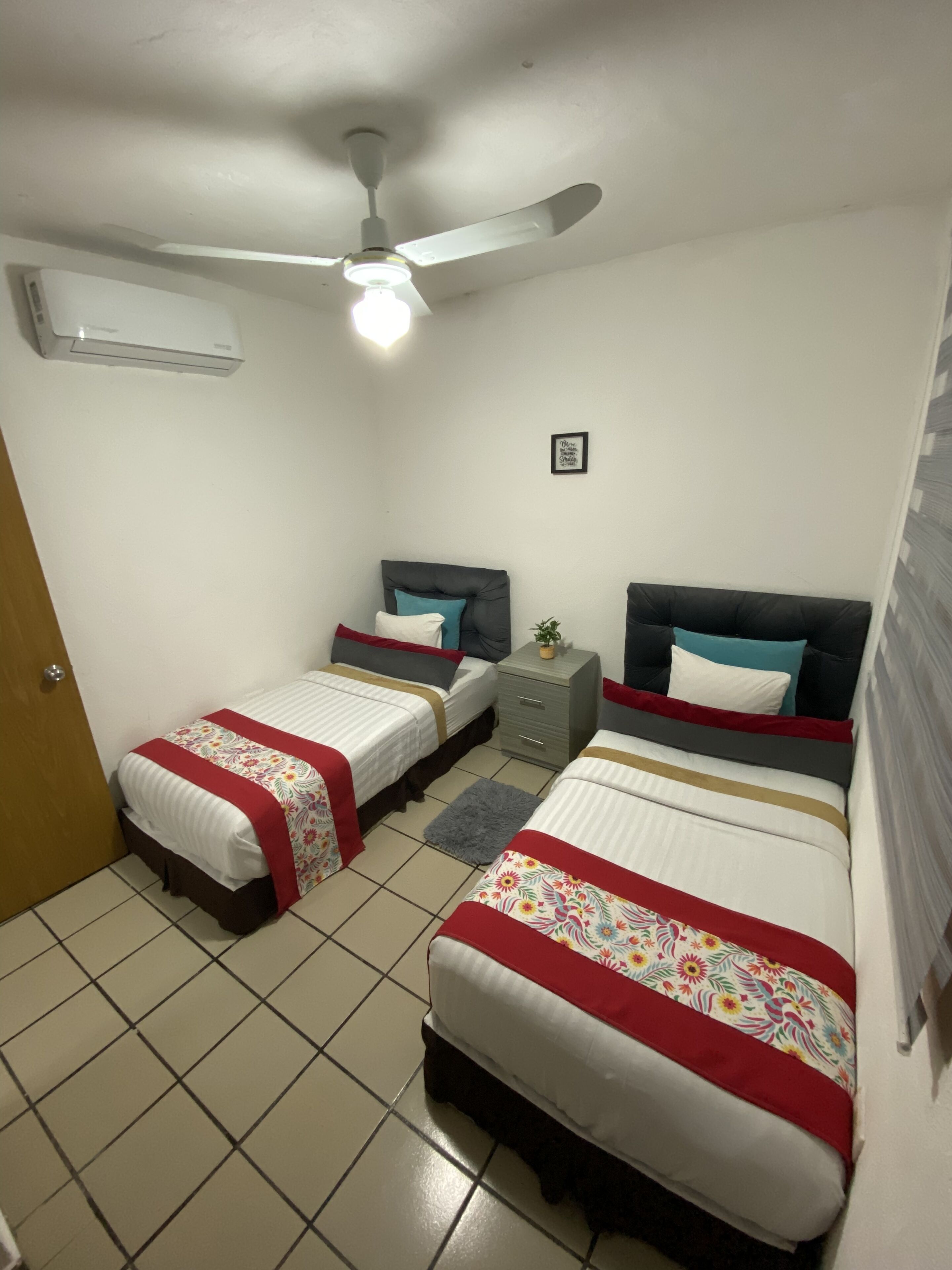 A small, clean room with two beds, a ceiling fan, an air conditioning unit, and a small bedside table with a plant.