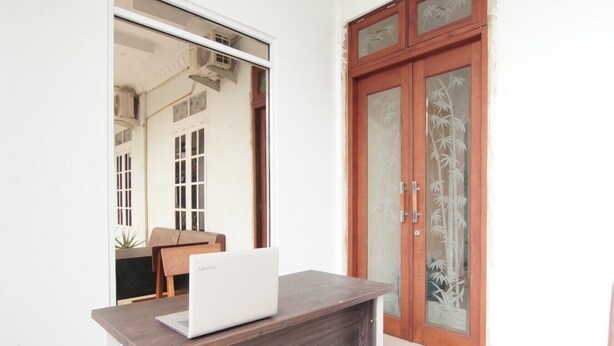 A room with a wooden desk, a laptop, a glass door with a bamboo pattern, and a view of a building with multiple windows and air conditioning units. - Hotel O Kng Homestay Syariah (Pekanbaru)