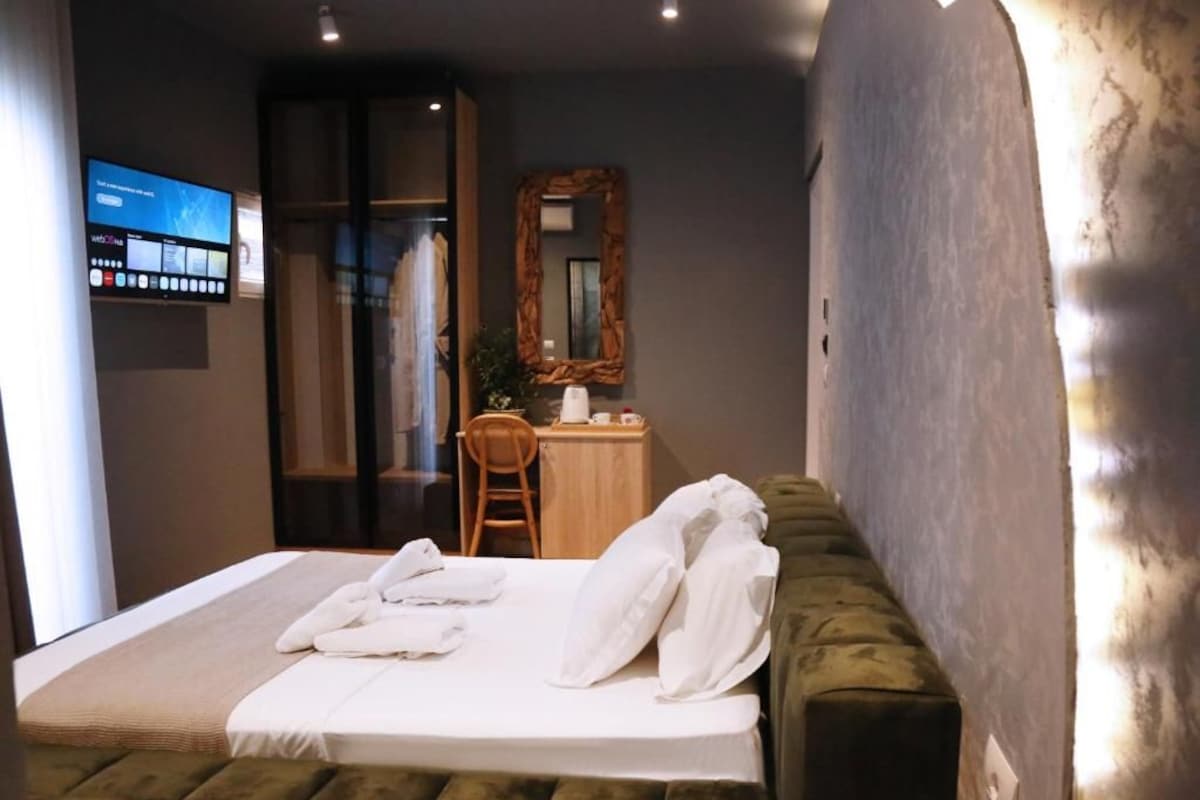 A modern hotel room with a large bed, a green upholstered headboard, a wooden desk, and a mirror on the wall.