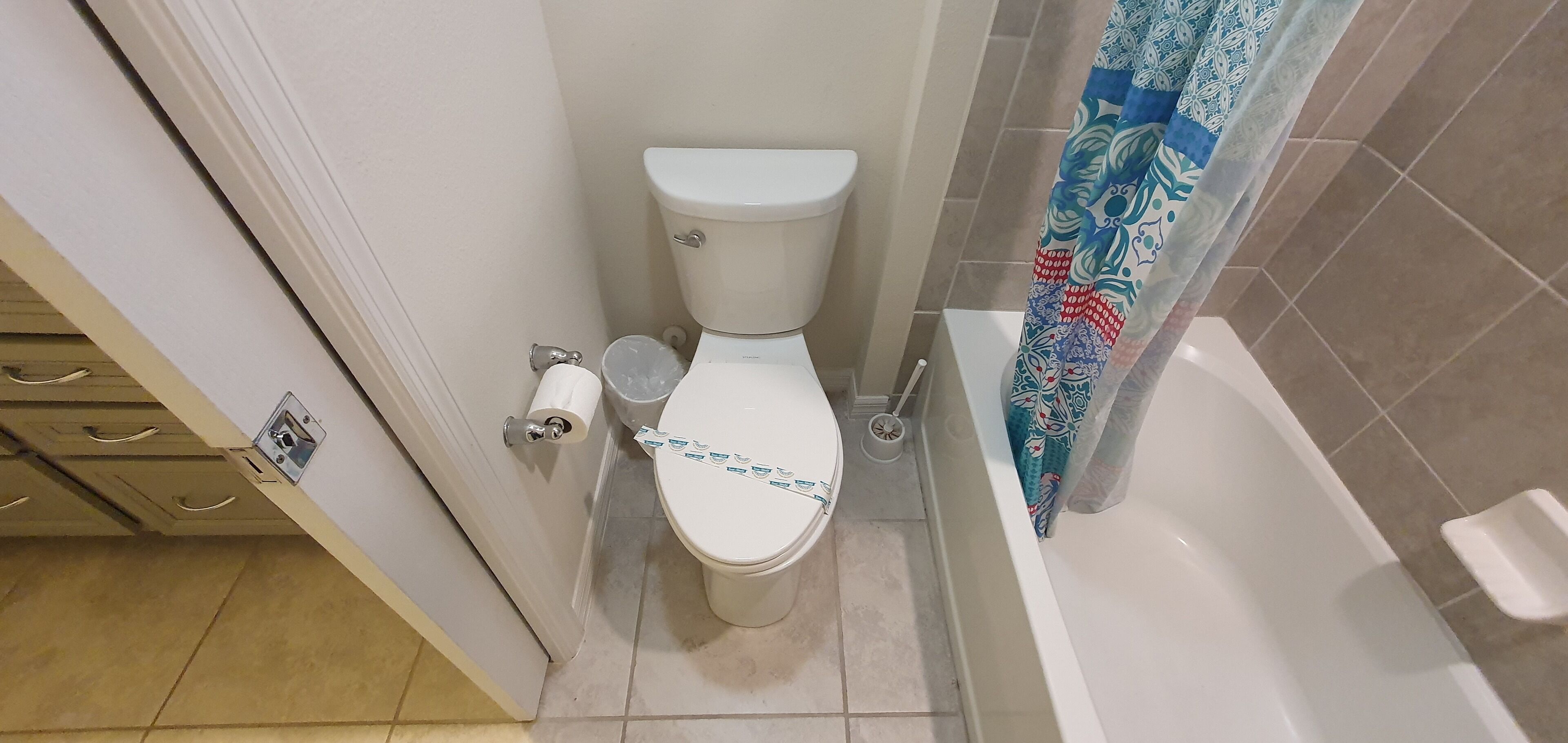 A bathroom with a toilet, a roll of toilet paper, a trash bin, and a bathtub.