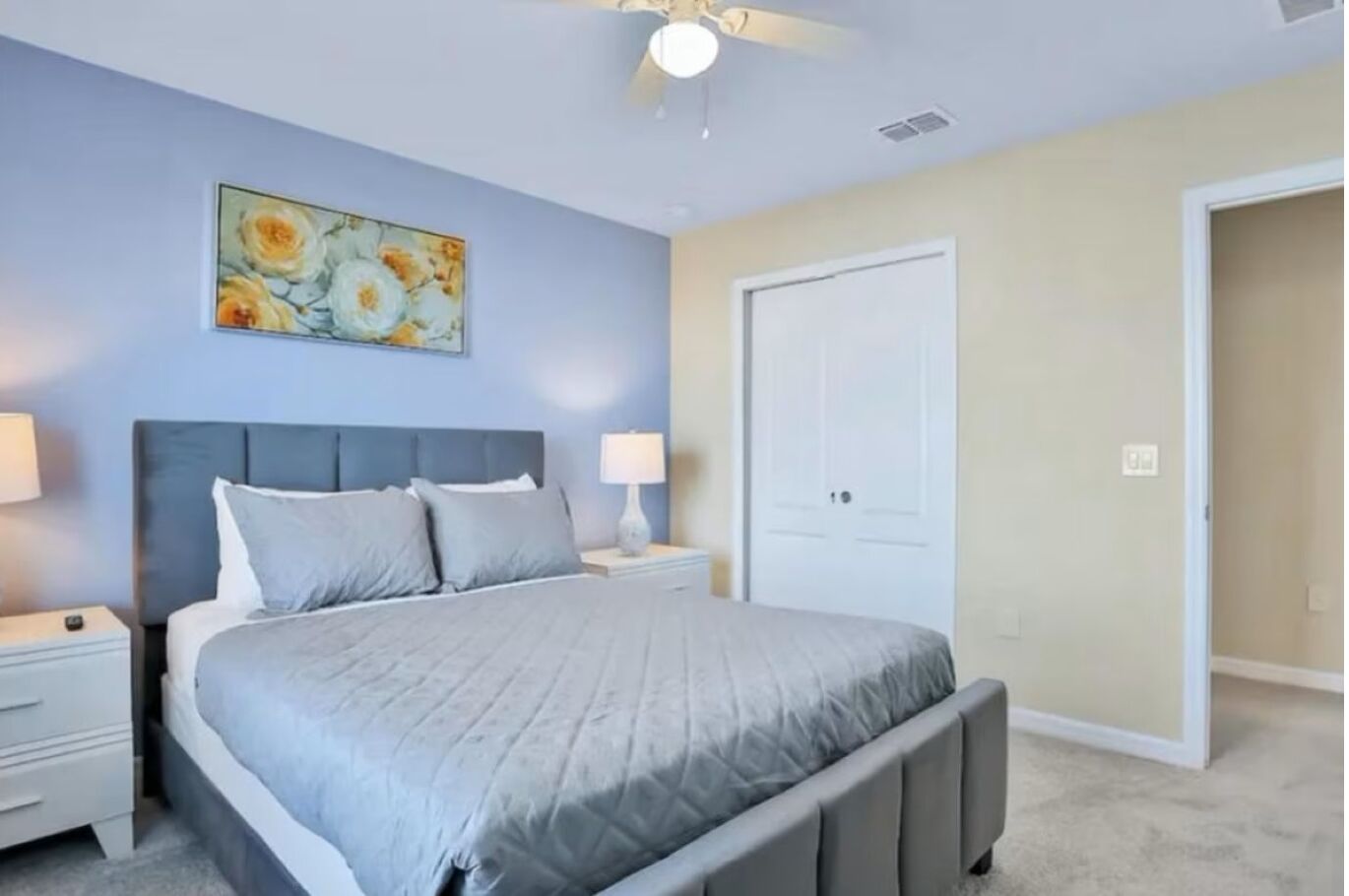 A bedroom with a bed, nightstands, a ceiling fan, a door, and a painting on the wall.