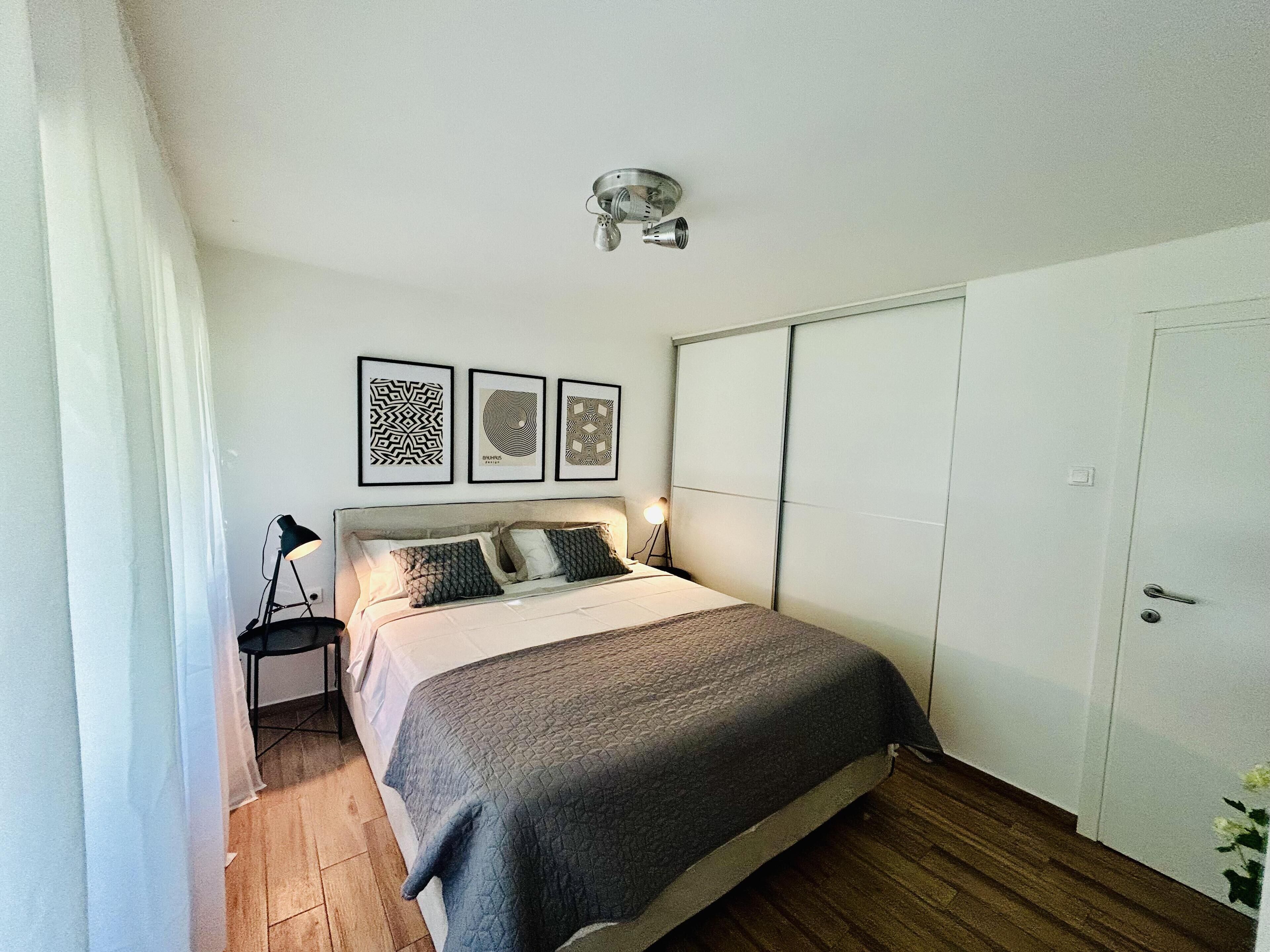 A bedroom with a bed, a bedside table, a lamp, a wardrobe, and three framed pictures on the wall.