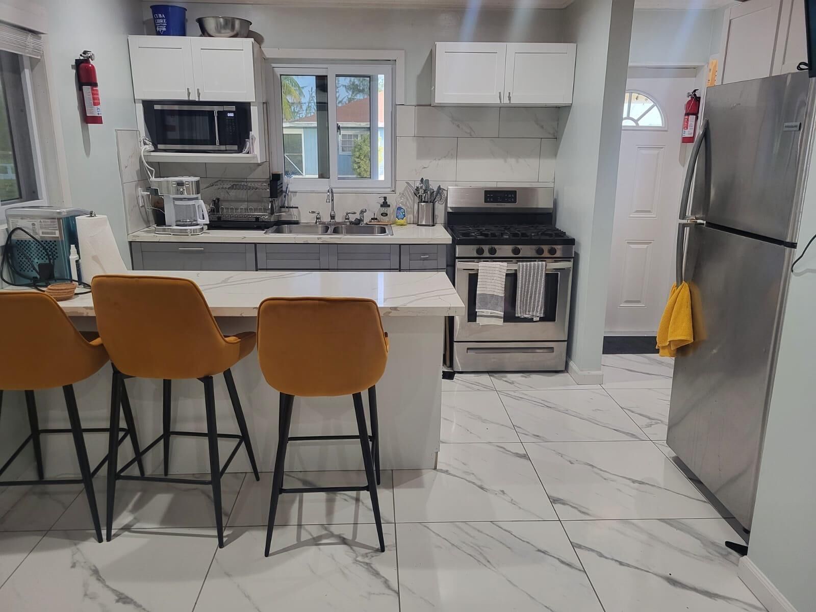 A modern kitchen with white cabinets, a stainless steel refrigerator, a gas stove, and a microwave. There are three orange chairs at the island.