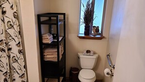 A small bathroom with a toilet, a shelf with towels, a window with a plant, and a shower curtain with a bird pattern. - Cabin #2 - Backyard Bunkhouse
 (Irwin)