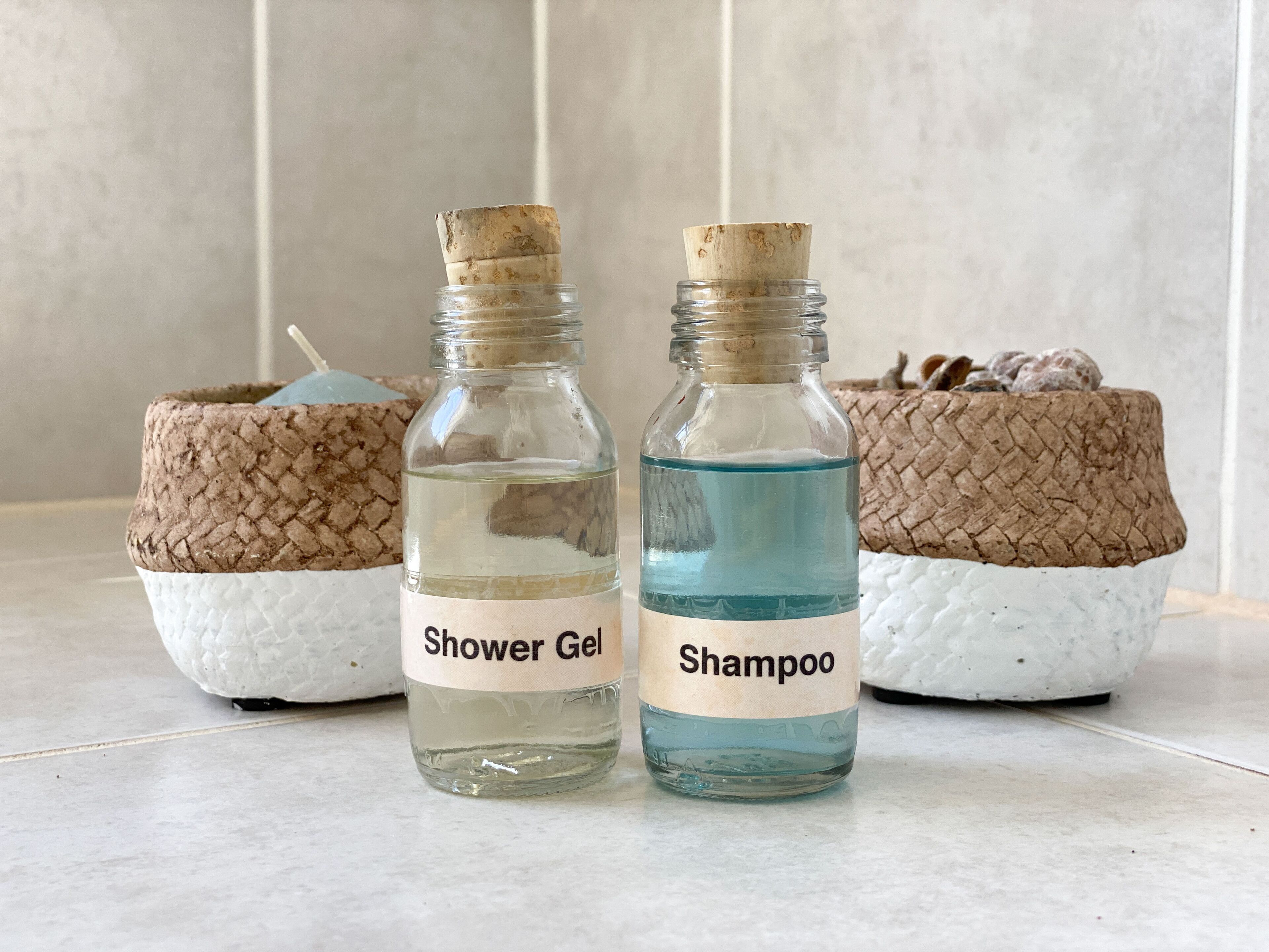 To glasflasker mærket 'Shower Gel' og 'Shampoo' med korkpropper, placeret på et flisegulv.