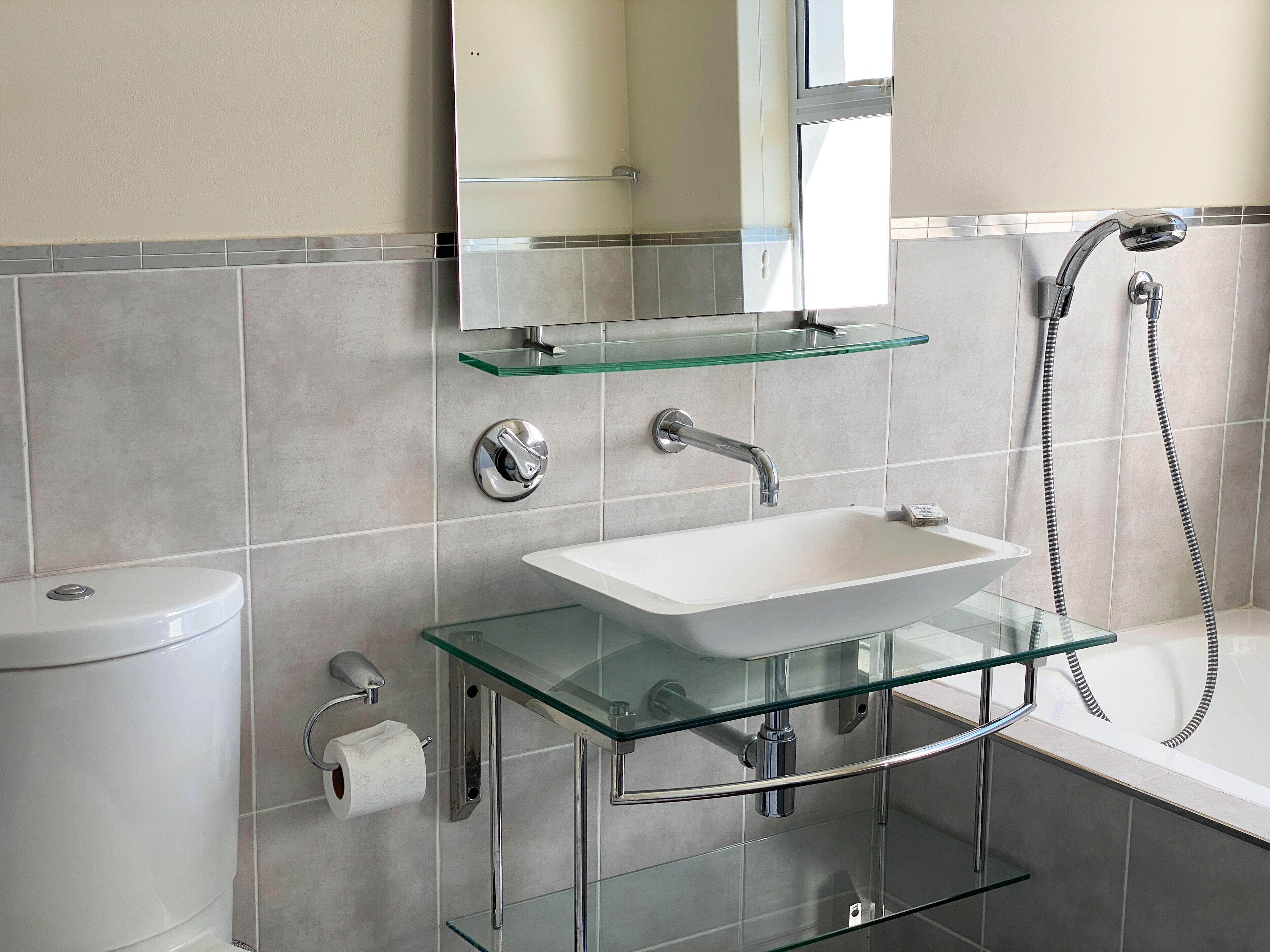 A modern bathroom with a glass sink, chrome fixtures, and a glass shelf above the toilet.