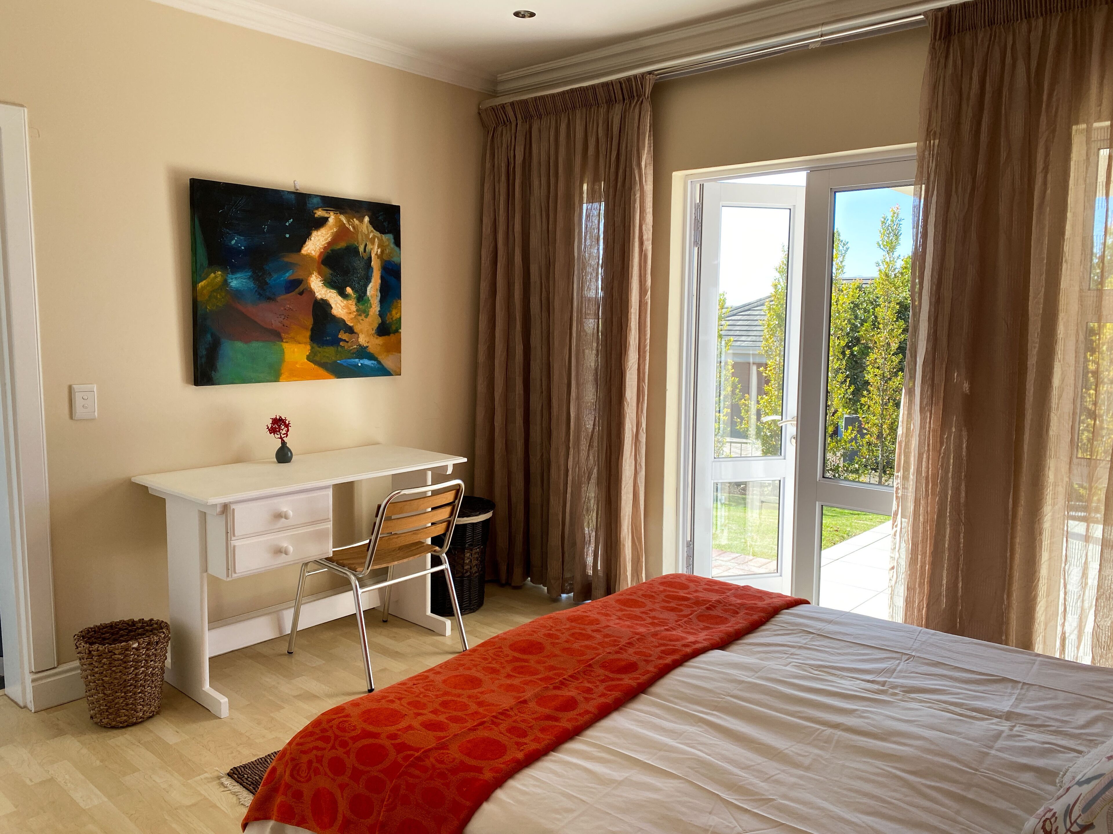 A bedroom with a bed, a desk, a chair, a painting, and a view through the window.