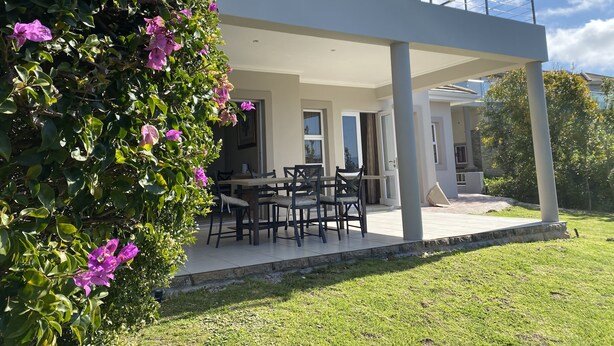 A modern house with a covered patio area, a dining table with chairs, and a lush green lawn.