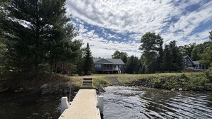 Une jetée en bois menant à une maison située au bord d’une étendue d’eau calme, entourée d’arbres.