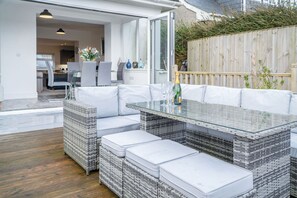 Ein moderner Essbereich im Freien mit einem Tisch mit Glasplatte, weißen Sitzmöglichkeiten mit Polsterung und einem Beistelltisch aus Rattan.