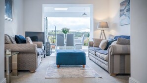 A modern living room with two sofas, a coffee table, and a TV. There is a balcony with a view of the countryside.