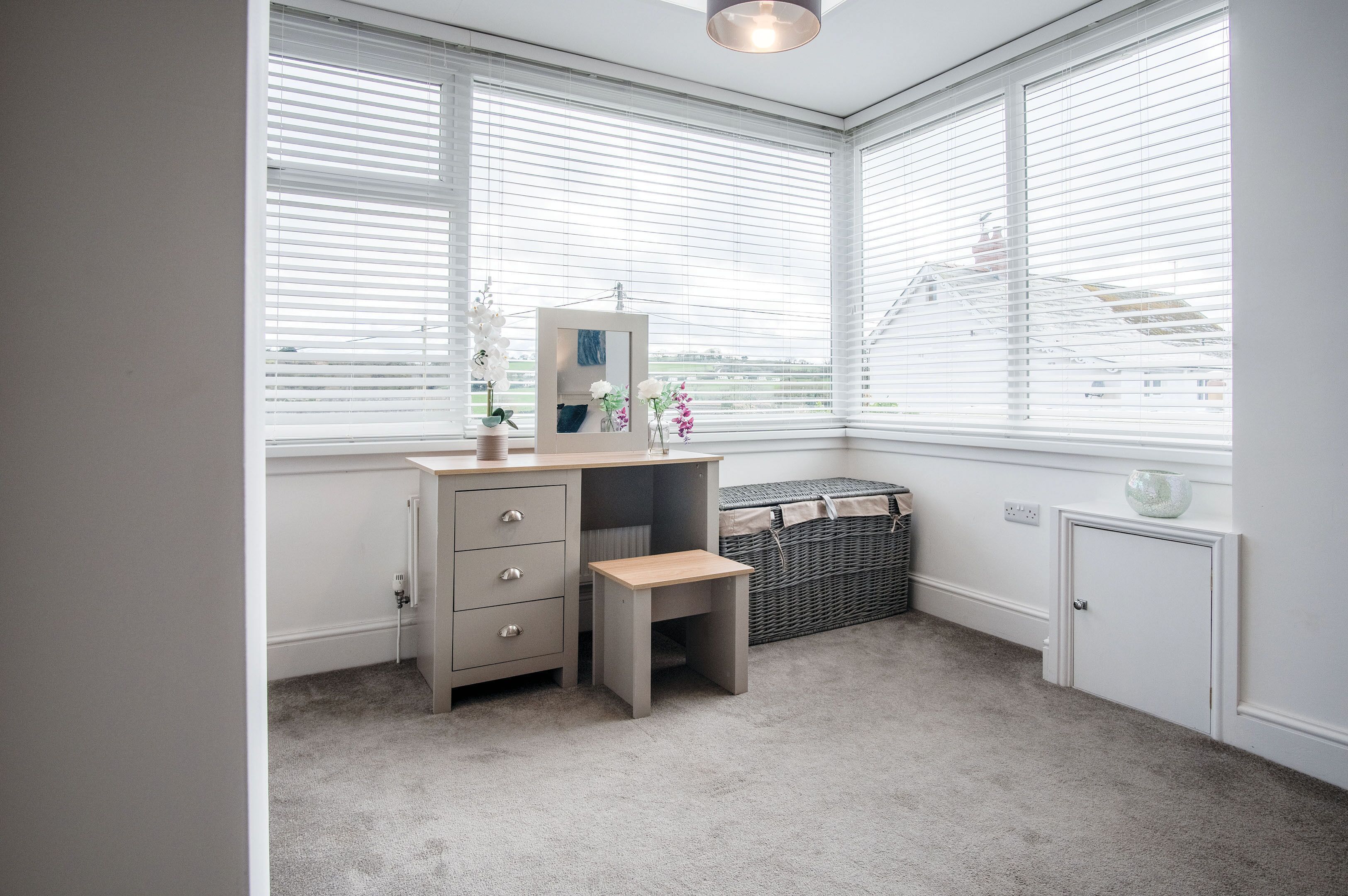 A corner bedroom with a desk, a small stool, a chest of drawers, a radiator, and a window with blinds.